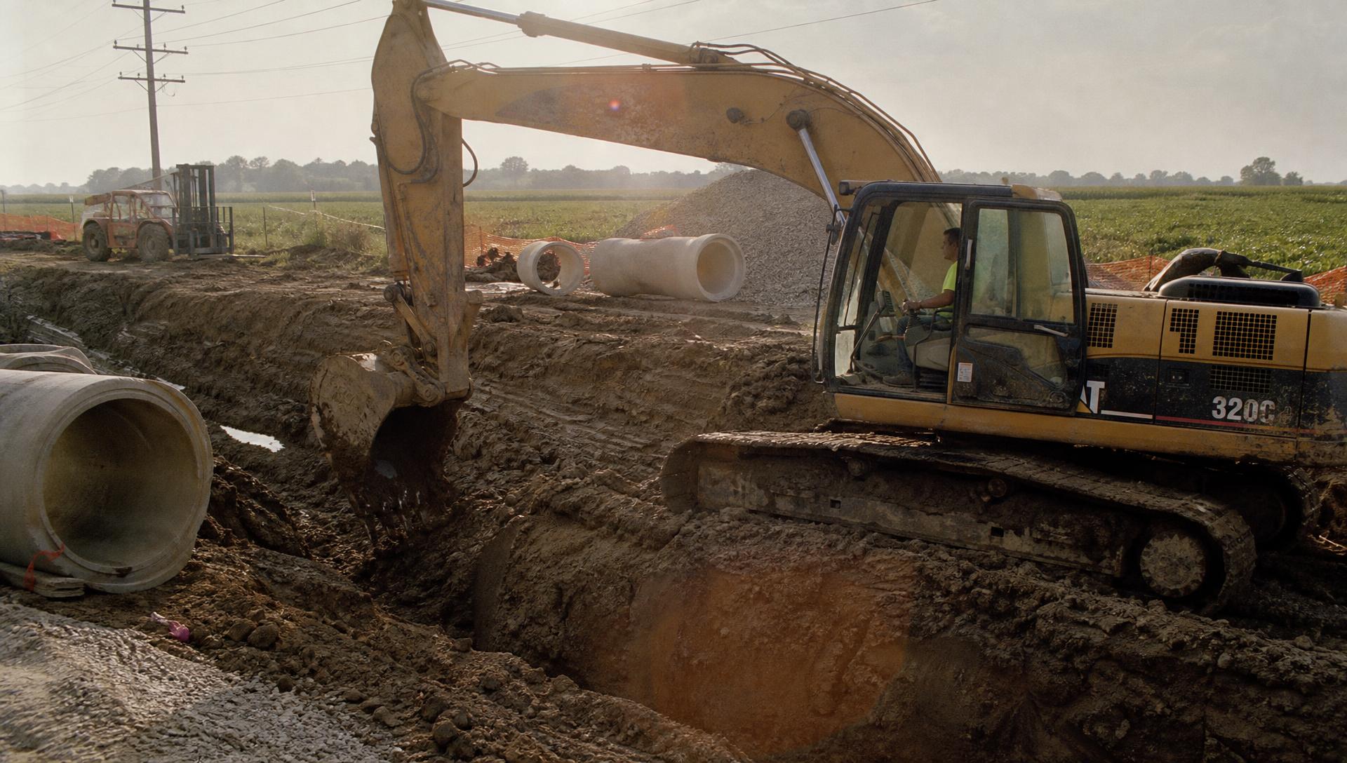 Excavator digging a utility trench at sunset with concrete pipe staged nearby